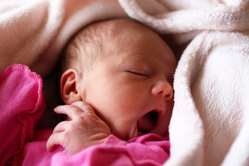 Cute newborn baby in pink baud with an open mouth on her bed under beige blanket.