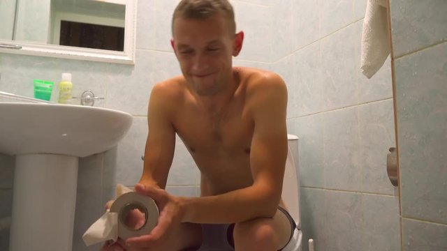 The person feels the pain of constipation in the toilet. A naked, naked man writhes on a toilet with toilet paper in his hands.