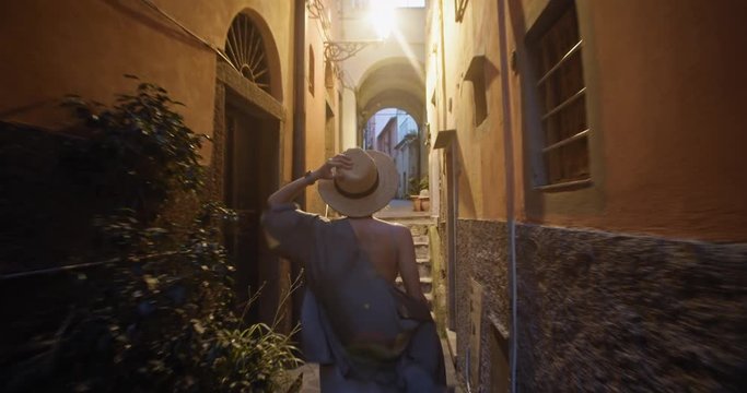 Stunning beauty blonde girl in sun hat walking in narrow street under sunlight. Back view of goddess woman exploring romantic Italian village of Riomaggiore.
