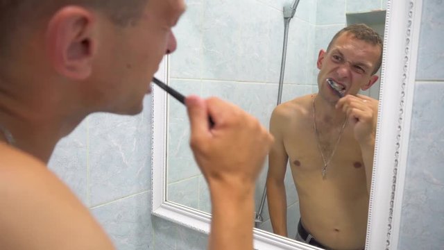 Man brushing his teeth getting ready in the morning doing hygiene routine looking in mirror of home bathroom using toothbrush in for clean dental oral care.