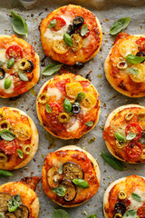 Small vegetarian pizzas with the addition various kinds of vegetables , mozzarella cheese and fresh basil on a on white baking paper, close-up, top view.