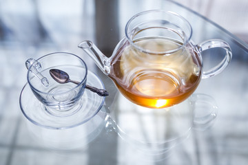 Transparent glass tea cup and pot with spoon on glass table in strong backlit