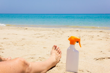 Sunscreen spray bottle on sand by legs of woman lying down on turquoise water empty beach. Summer holidays, travel vacation, skin protection concepts