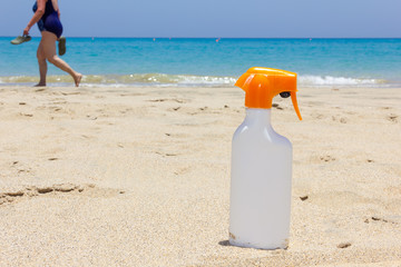 Fototapeta na wymiar Sunblock white bottle on sand as woman passes by shore on turquoise water beach. Sunscreen spray, skin care protection, summer vacation concepts