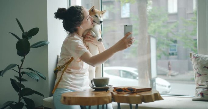 Happy lady is taking silfie with adorable dog using smartphone camera having fun sitting on window sill in cafe holding device wearing wireless earphones. Youth and animals concept.