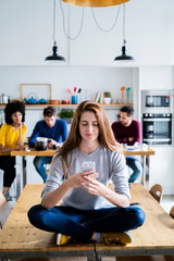 Woman with cell phone sitting on dining table at home with friends in background