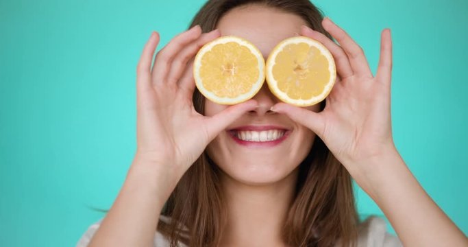 Funny portrait of young attractive brunette on turquoise backgroung is taking splitted halfs of yellow lemon and closes his eyes, smiling and laughing. Slow motion.