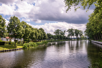 Obraz premium Sluis, the Netherlands - June 16, 2019: Reflecting water of canal to Damme in Belgium, with curtains of green trees on sides, under a storm approaching cloudscape.