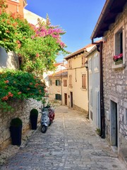 A beautiful classic narrow cobblestone European street with colourful flowers and a motorcycle parked.  There are no people or cars.  It is a sunny day in Rovinj, Croatia.
