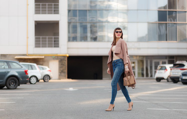 Portrait of beautiful brunette young woman in nice brown beige coat, denim jeans and sunglasses. Genuine leather bag, high heels shoes. Spring autumn fashion trendy photo of lady on urban background.