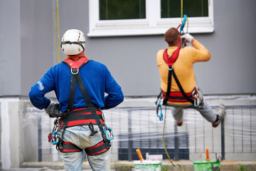 Professional industrial alpinist with rigging equipment, hardhat and safety harness from the back. Painter hanging on cable with paint buckets, industrial climber repairing house facade
