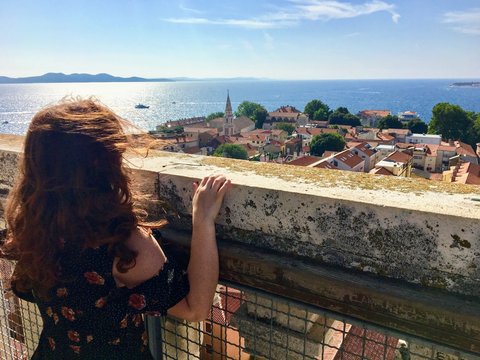 A young female tourist at the top of the bell tower in the old town of Zadar, Croatia, looking out at the beautiful town and the adriatic sea in the distance on a beautiful summer day.