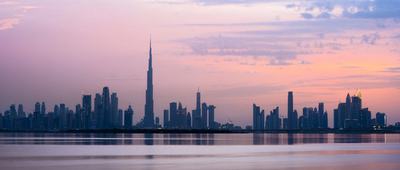 Atemberaubende Aussicht auf die Skyline von Dubai bei Sonnenuntergang mit dem prächtigen Burj Khalifa und vielen anderen Gebäuden und Wolkenkratzern. Seidig glattes Wasser, das im Vordergrund fließt.