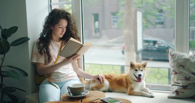 Female student is reading book and stroking pet dog sitting on window sill in cafe enjoying hobby and leisure time. Literature, animals and youth lifestyle concept.