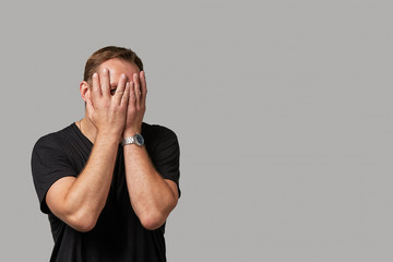 A man in casual clothing hides his face with his hands on a gray background. Emotional, brave face. Isolated on white background Copy space Advertising and commercial design.