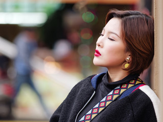 Beautiful young brunette woman in black coat lean against wall with eyes closed. Outdoor fashion portrait of glamour Chinese stylish lady in street. Emotions, people, beauty and lifestyle concept.