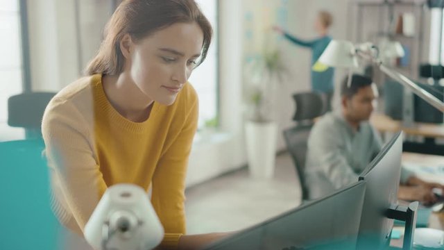 Beautiful and Creative Young Woman Sitting at Her Desk Using Laptop Computer. In the Background Bright Office where Diverse Team of Young Professionals Work 