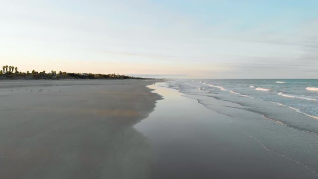 aerial view of a sunrise on the beach of rota, cadiz, you can see 3 people sitting on the sand and another person walking, in the fodo you see houses and trees. The sun is at the bottom