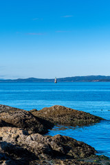 Rocky coastal landscape along the ocean with floating lighthouse in the background. Clear sunny autumn day with mountains in the background beyond the deep blue sea water.