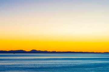 Obraz premium Beautiful twilight with golden sunset/sunrise over the ocean behind mountain silhouette. Orange yellow blue skies over water as the sun sets/rises. Harmony and beauty in nature. Scenic peaceful view.