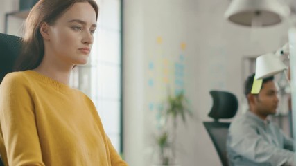 Portrait of Beautiful and Creative Young Woman Sitting at Her Desk Using Laptop Computer. In the Background Bright Office where Diverse Team of Young Professionals Work - Powered by Adobe