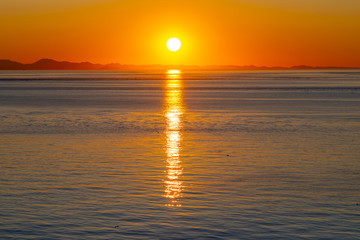 Beautiful golden sunset/sunrise over the sea behind mountains in background. Light reflection on the water as the sun sets/rises. Harmony and beauty in nature. Scenic peaceful ocean view.