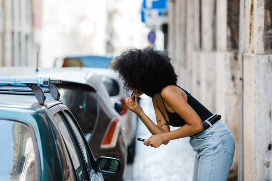 Side view of young ethnic woman looking at car window and doing makeup while standing on urban background