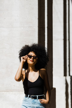 Young ethnic woman in jeans and tank top leaning on wall and smiling at camera outdoors