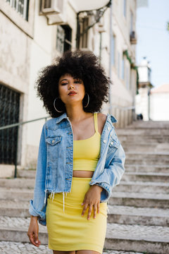 Beautiful African American woman in yellow suit standing on stairs and looking at camera on urban background