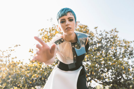 From below young lady with short blue hair and in futuristic dress bending forward and trying to touch camera on blurred background of tree on sunny day in park