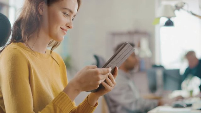 Talented Female Designer Sitting at Her Desk She's Holding and Using Touch Screen Digital Tablet Computer. Bright Office where Diverse Team of Young Professionals Work