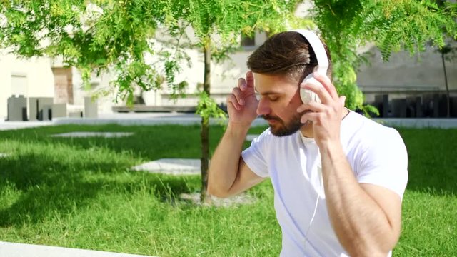 Smiling handsome young man listening music from his smartphone in trendy white headphones near nice green yard
