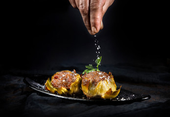 Crop hand of person sprinkling tasty stuffed artichokes with salt on black background
