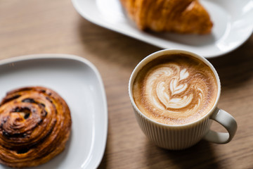 Cappuccino with a pattern of leaves from cinnamon on milk foam with the homemade cinnamon bun and freshly baked croissant on the wooden table.