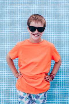 Smiling male kid in bright orange T-shirt and sunglasses standing on background of tiled blue wall