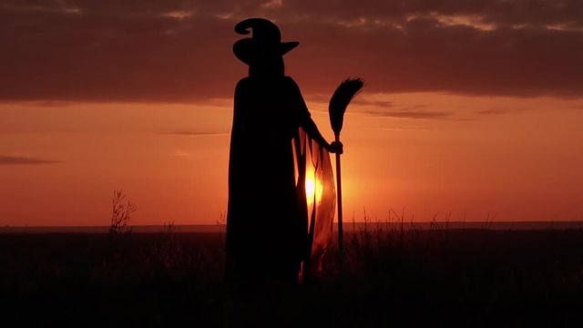 witch holding broom looking on the amazing red sky with orient sun standing on the hill
