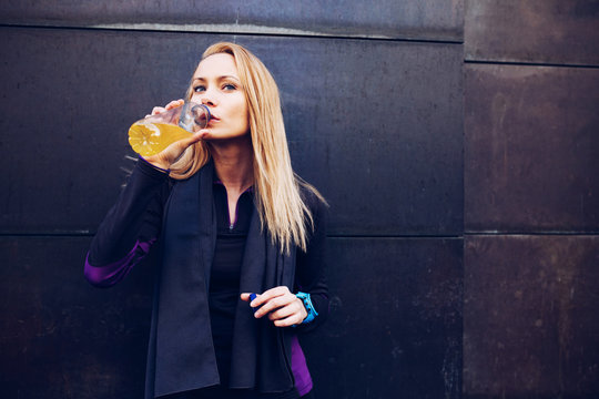 Young blonde caucasian woman drinking isotonic drink while having a rest from a running session leaning in a wall