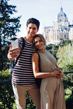 Young handsome man taking photo with girlfriend while walking in beautiful garden on background of historical building