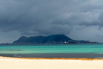 Colorful view from the beach de Getares: white sand contrast with azure sea and dark sky. Fluffy clouds over the Gibraltar. Impressive ships are constantly in the Bay. Algeciras, Andalusia, Spain.