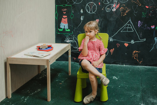 Adorable child thinking in light green chair with crossed legs in creative room at table