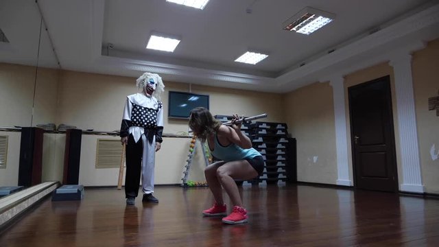 pretty lady in pink sneakers squats with silver metal bar near scary clown with mask in large modern gym