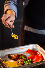 Chief cook is decorating a meat dish with corn at kitchen background. Vertical image.
