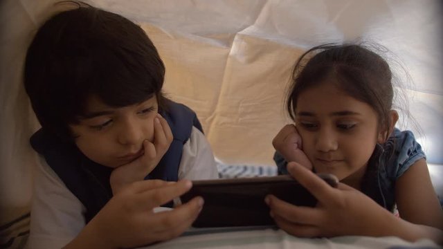 Indian siblings using tablet pc under the blanket at night - Technology invasion . Cute little kids under the blanket and watching cartoon movie on their tablet - Use of technology in personal lives
