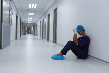 Fototapeta premium Tensed female surgeon with hands on forehead sitting on floor in the corridor at hospital