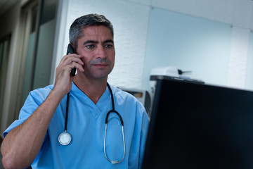 Male surgeon talking on mobile phone working on computer in hospital