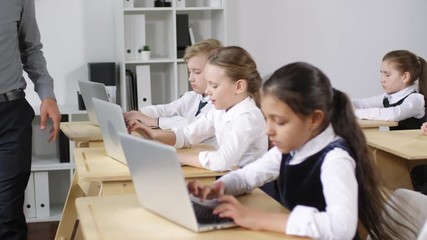 Medium shot of diligent preteen schoolchildren sitting at desks at computer education lesson and working with laptops on assignment, and male teacher pacing around and checking on them - Powered by Adobe
