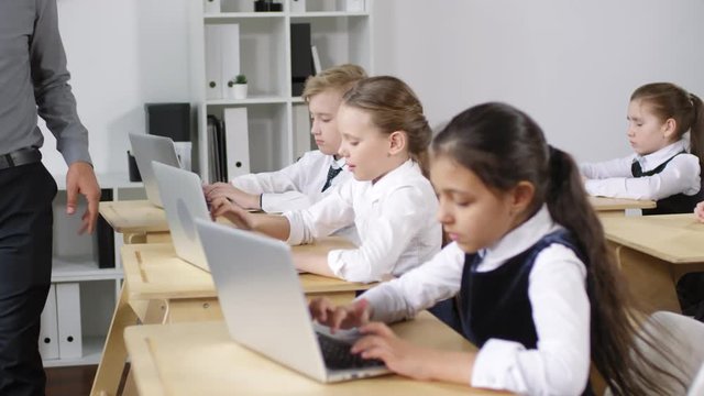 Medium shot of diligent preteen schoolchildren sitting at desks at computer education lesson and working with laptops on assignment, and male teacher pacing around and checking on them