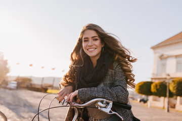 Pretty european woman with gorgeous curly hair posing in cold autumn day in city square. Elegant brunette lady with black scarf driving bicycle around city.