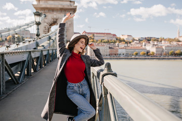 Pretty young woman in blue jeans and long coat dancing on bridge near triumphal arch. Outdoor...