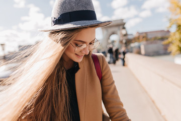 Romantic fair-haired girl in beige outfit looking down with smile. Outdoor photo of graceful woman in gray hat decorated with black ribbon enjoying sunny day in autumn.